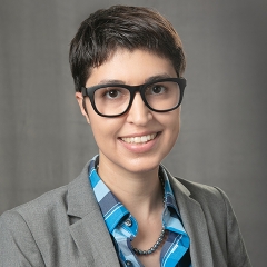 Harlow Loch Ramirez in black glasses, a blue shirt, and a grey overcoat, smiling at the camera in front of a grey background.